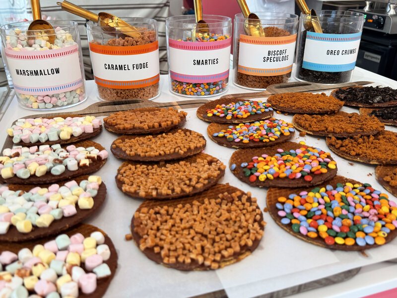 Five labelled jars of toppings with gold scoops above a tray of finished chocolate stroopwafels