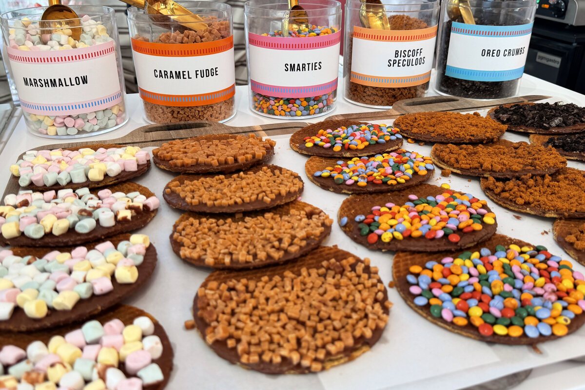 Five labelled jars of toppings on the counter, marshmallow, caramel fudge, smarties, Biscoff speculoos and Oreo crumbs, with gold scoops, above a tray of finished chocolate stroopwafels