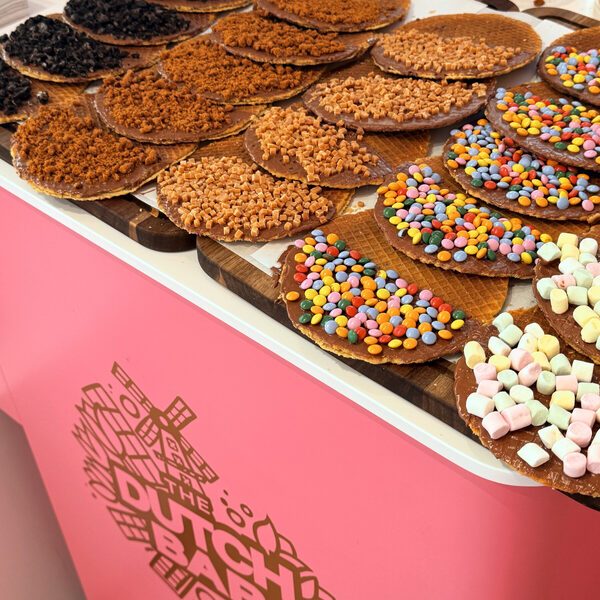 Wooden boards of chocolate-dipped stroopwafels with different toppings on the pink counter, the gold Dutch Bar windmill logo embossed on the counter front