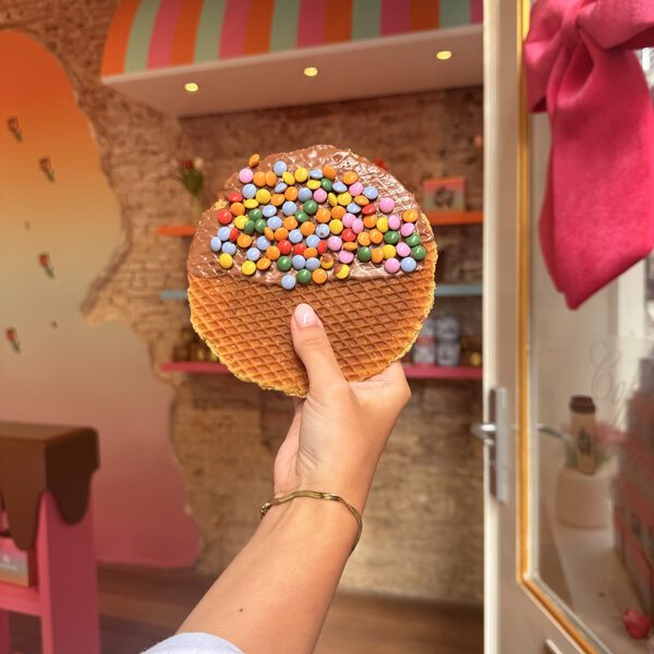 A smarties stroopwafel held in the doorway, with the striped awning, bare brick wall and pink chocolate-drip shelving inside