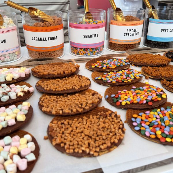 Five labelled jars of toppings on the counter, marshmallow, caramel fudge, smarties, Biscoff speculoos and Oreo crumbs, with gold scoops, above a tray of finished chocolate stroopwafels