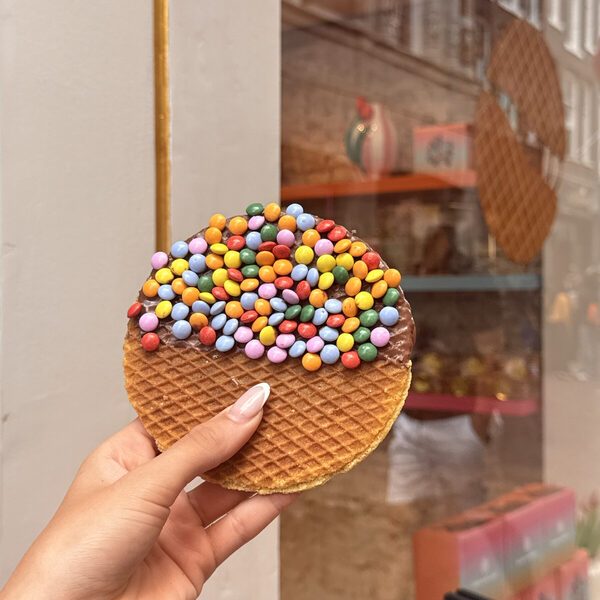 A giant stroopwafel covered in milk chocolate and coloured chocolate beans, held in front of the shop window