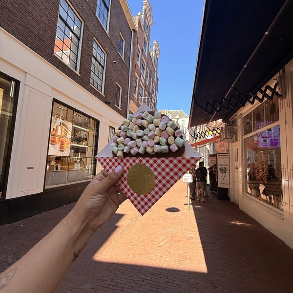A marshmallow stroopwafel held up in the Voetboogstraat under a blue sky, canal houses ahead and the queue at Vleminckx on the right